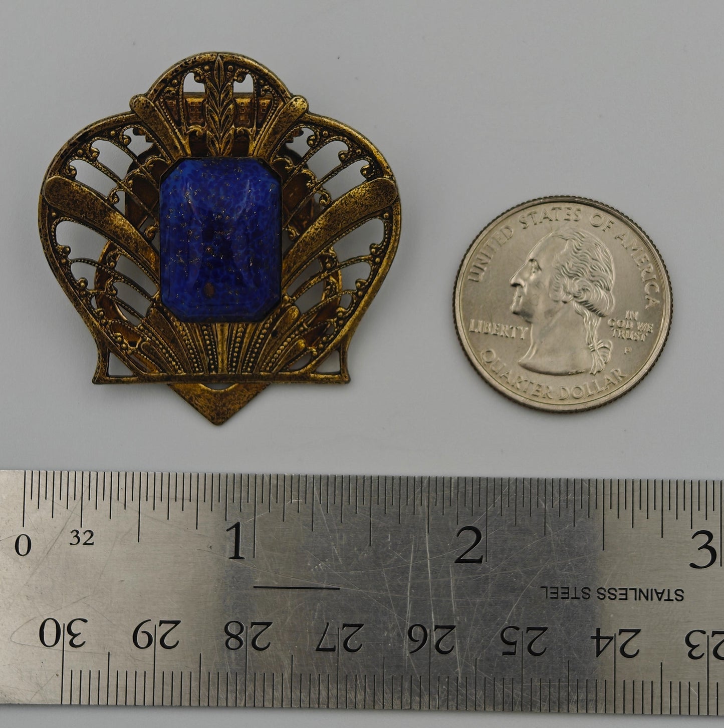 Decorative brooch with blue center and gold frame next to a coin and ruler for scale on a white background