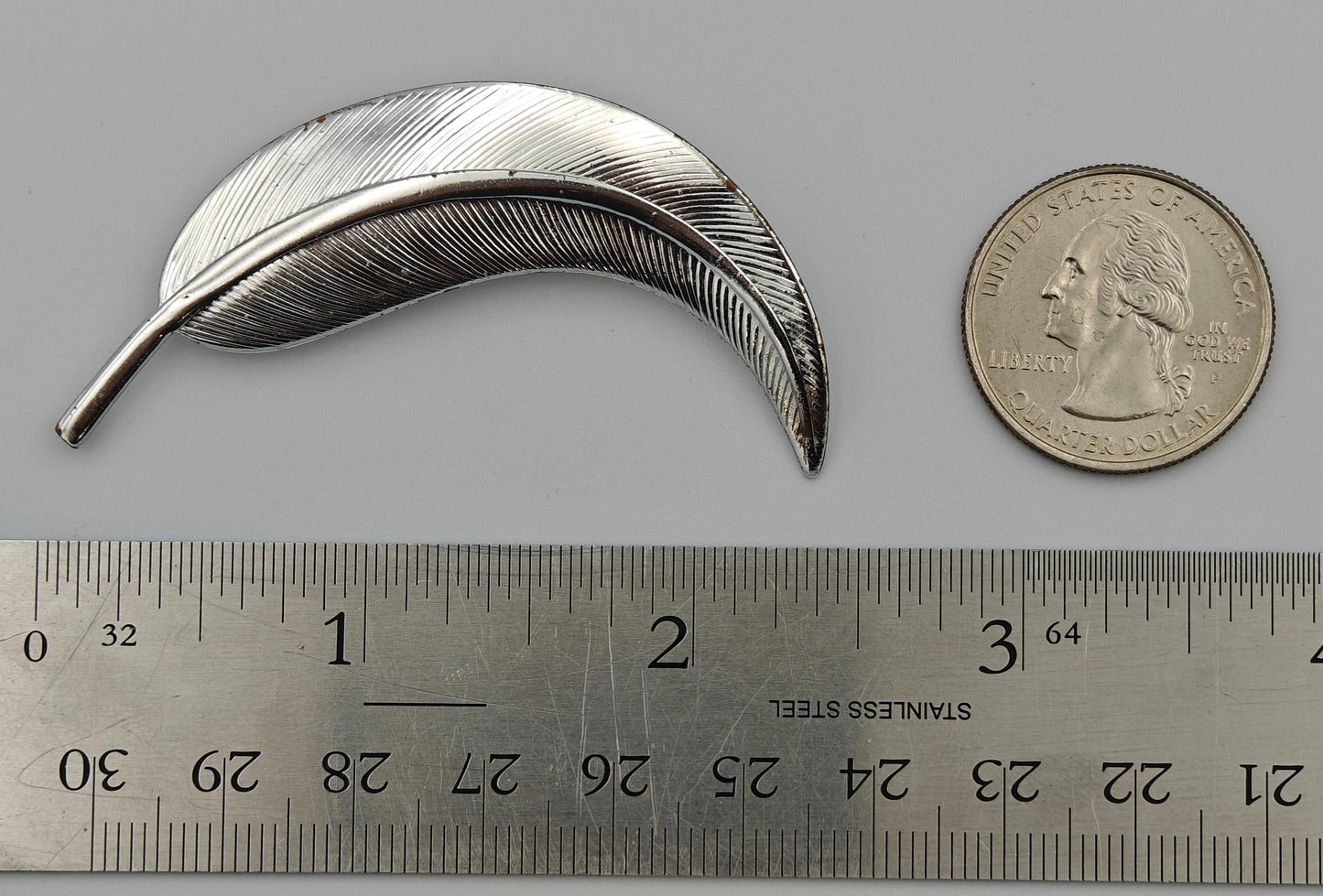 Silver feather-shaped brooch next to a ruler and a coin on a gray background