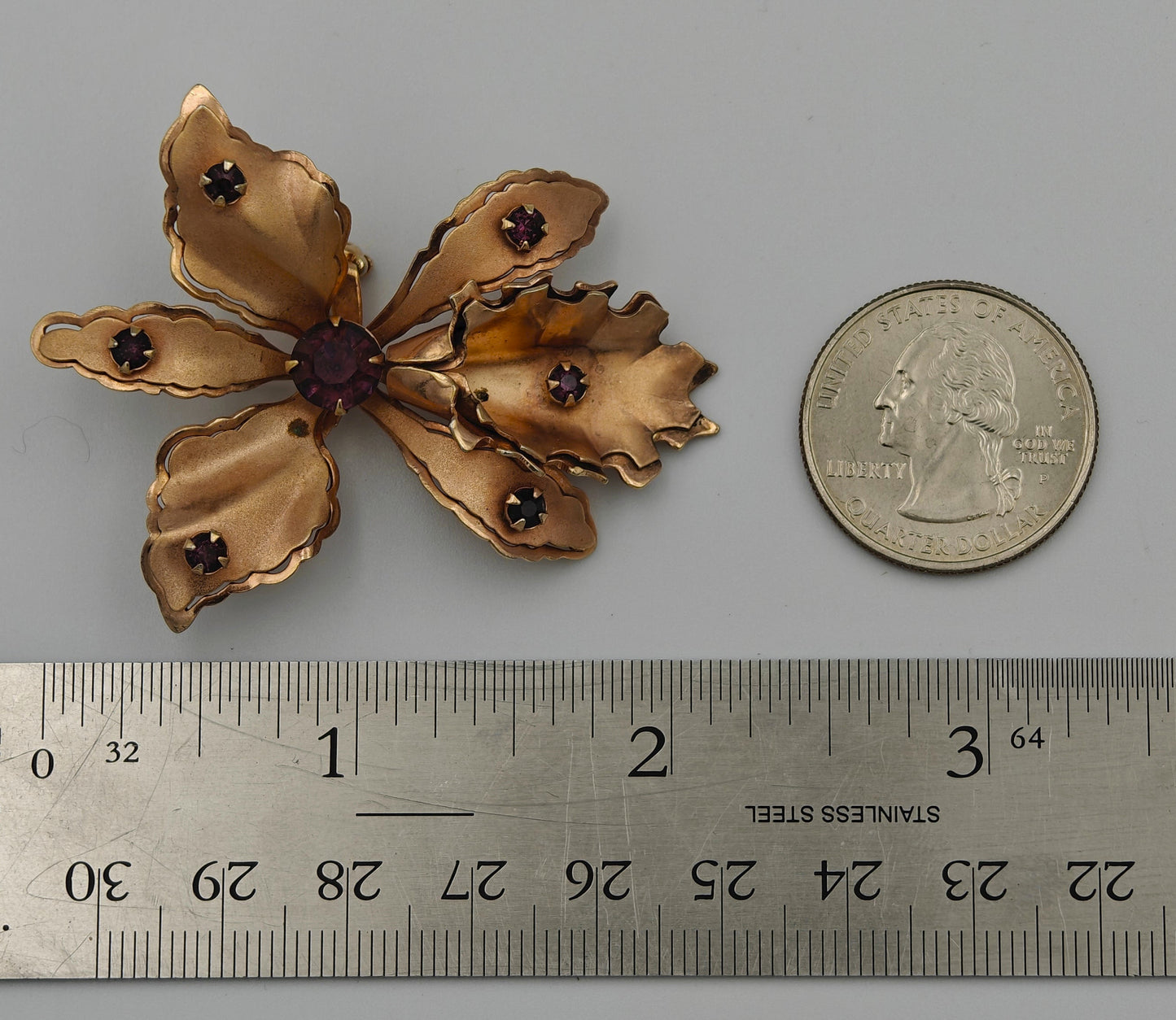 Gold flower-shaped brooch with dark gemstones next to a ruler and quarter for scale on a gray background