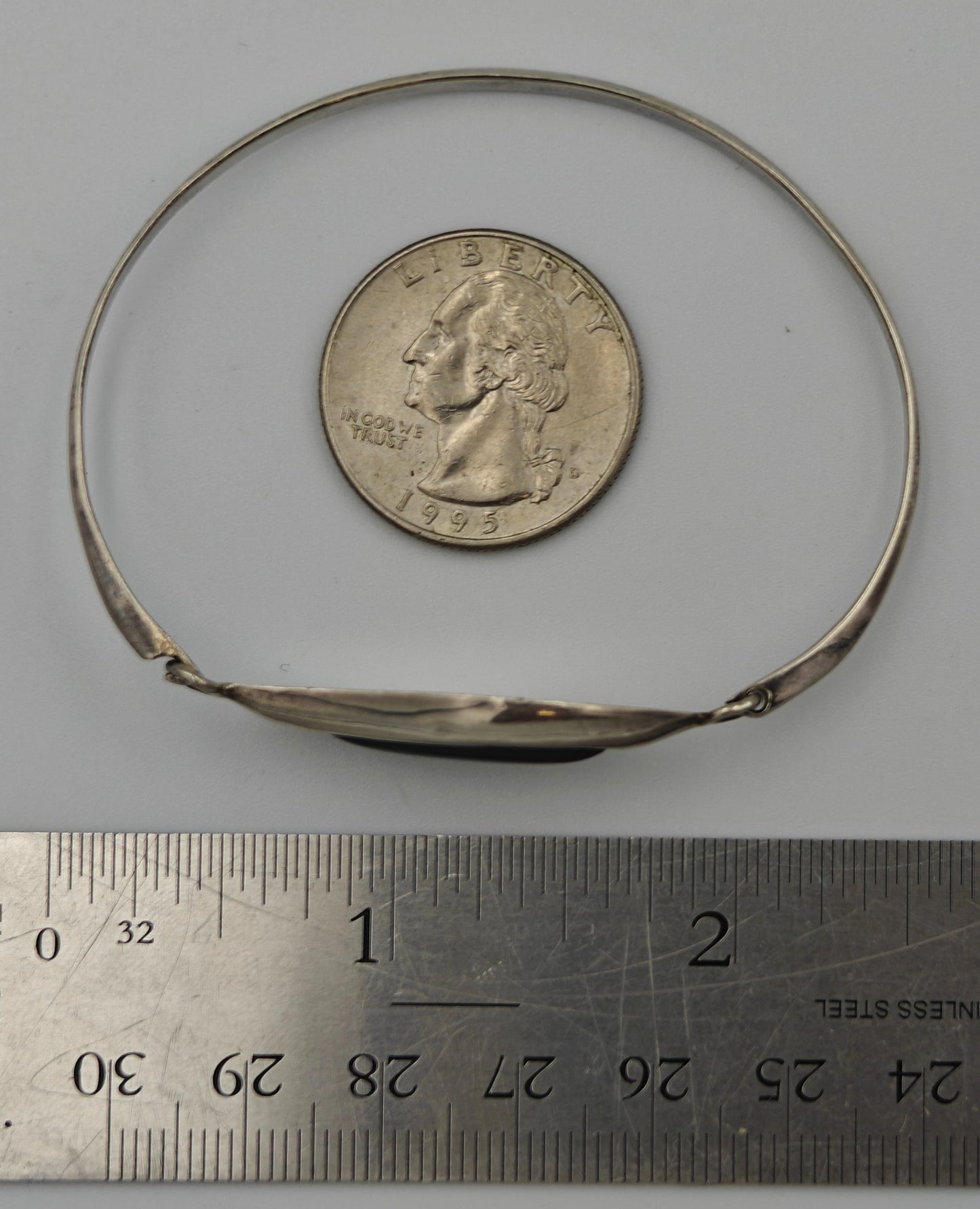 Silver bracelet next to quarter and ruler for scale on a white background.
