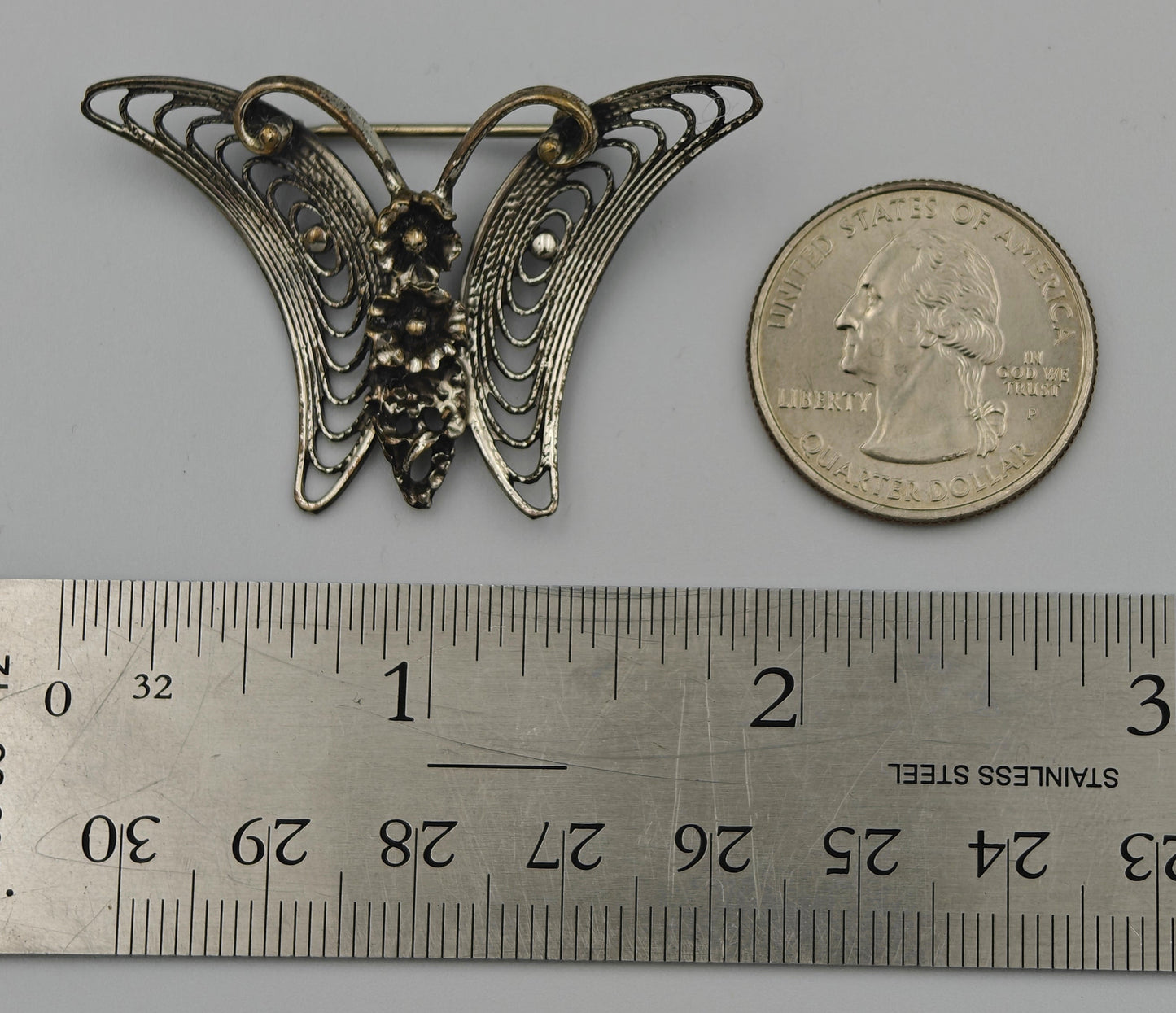 Butterfly-shaped brooch next to a U.S. quarter dollar on a gray background with a ruler for scale.