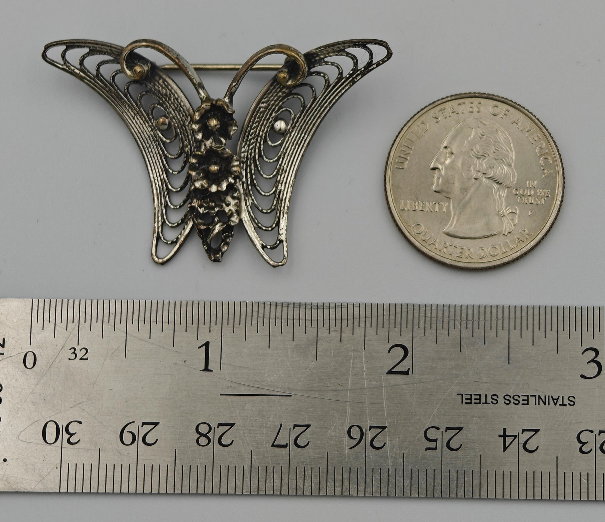Butterfly-shaped brooch next to a U.S. quarter dollar on a gray background with a ruler for scale.