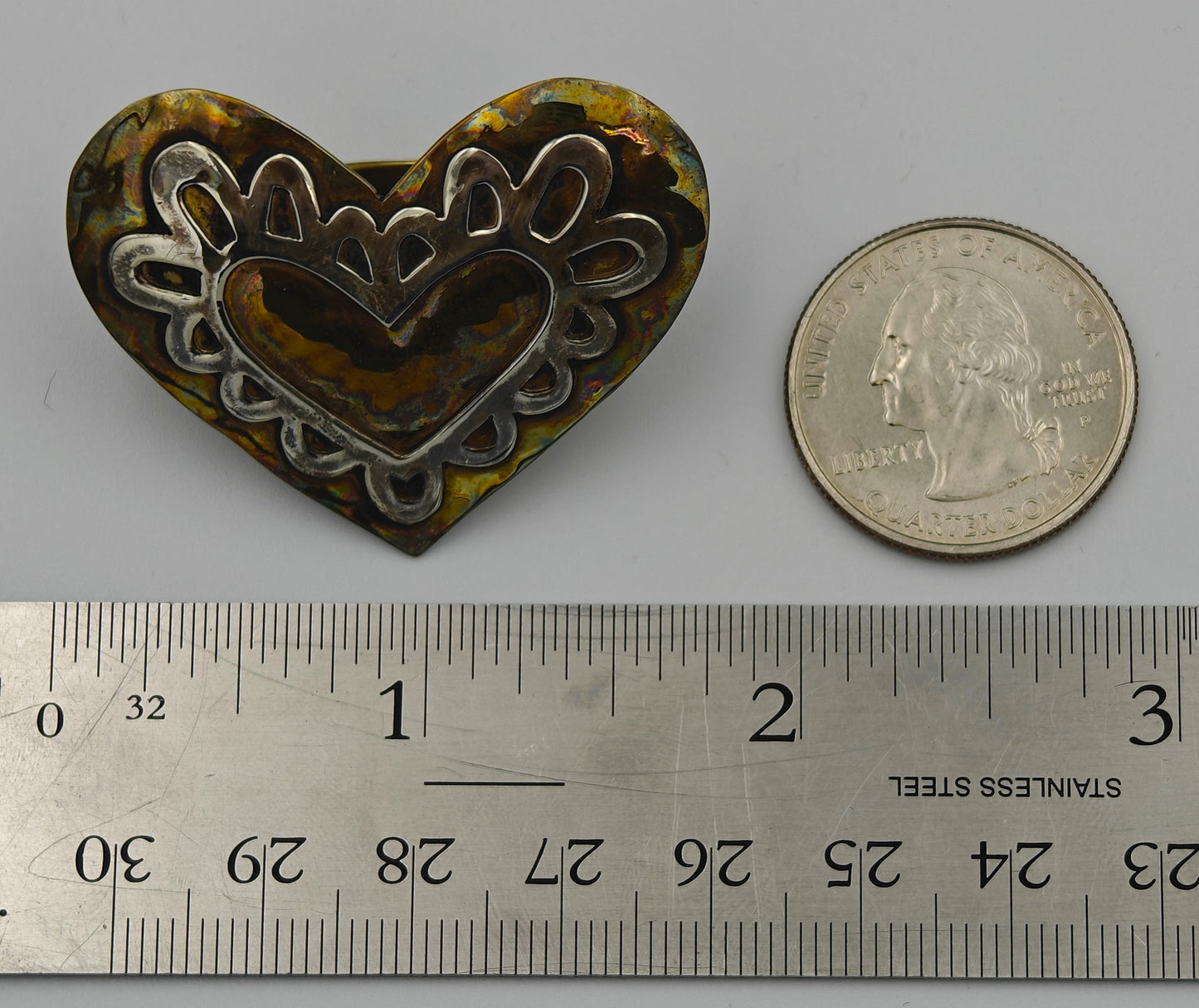 Heart-shaped metal scarf tie next to a ruler and coin on a gray background