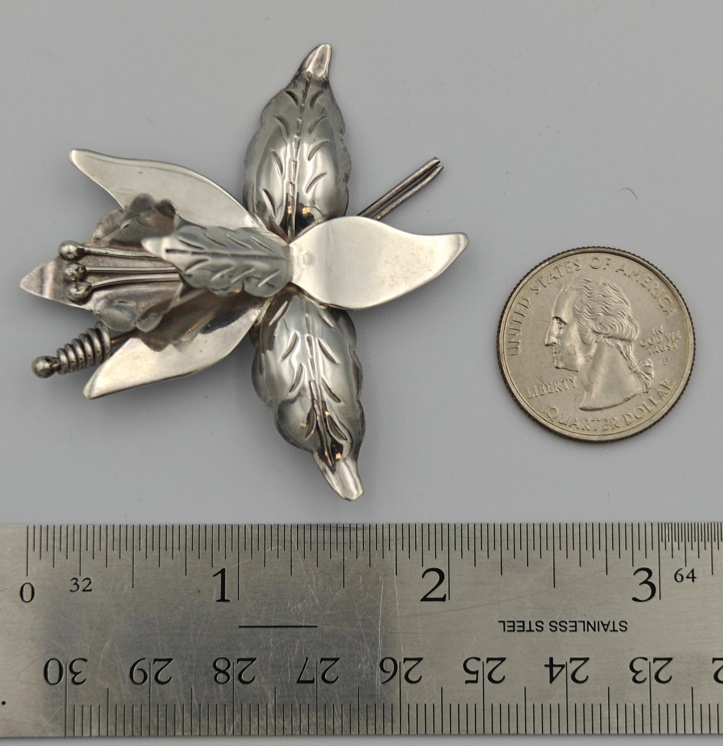 Silver flower-shaped brooch next to a ruler and coin on a gray background